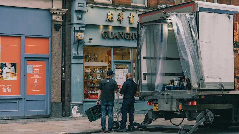 Two men from Man and Van Kentish Town are preparing for a home relocation outside a shop with Chinese signage that reads 'GUANGH' and includes Chinese characters. One man, wearing a black cap and dark jacket, is holding a black plastic crate, while the other, dressed in a dark hoodie, is pushing a small hand truck. They are standing on a pavement next to a large, open-moving truck with a transparent plastic curtain on the side, parked close to the shop entrance. Inside the truck, visible packing materials and packing crates indicate ongoing packing and furniture transport activities. The shop’s display window and doorway are reflected in the truck’s side. The scene captures the process of loading or unloading household items, with the backdrop of an urban building featuring traditional Chinese characters and signage, suggesting that the moving process is part of a professional relocation service provided by Man and Van Kentish Town, supporting efficient and carefully managed house removals in the area.