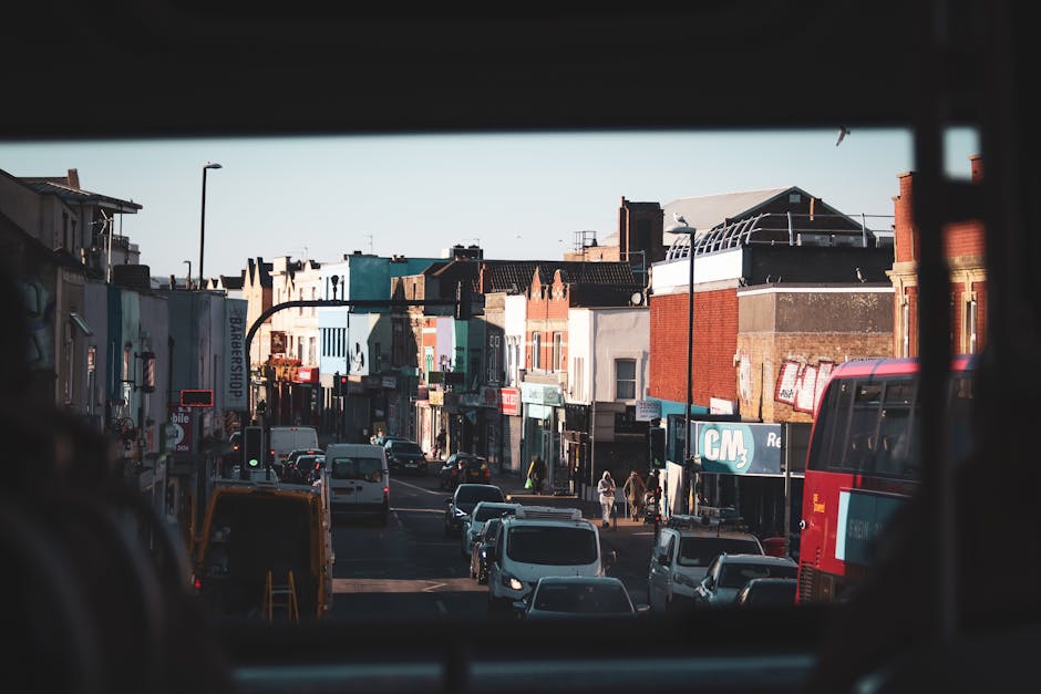 View of a busy street in Kentish Town captured from inside a vehicle, showing a variety of parked and moving cars, including white vans and a red double-decker bus on the side of the road. The street is lined with small shops, cafes, and commercial buildings with colorful facades and signage, some with awnings. Pedestrians are walking along the sidewalk, with a few crossing the street at the intersection, and streetlights are visible alongside the pavement. The image is taken in daylight, with clear weather and bright natural light illuminating the scene. This urban environment reflects typical city traffic and retail activity, relevant to house removals and moving logistics services offered by Man and Van Kentish Town, including the effort involved in furniture transport and home relocation processes.