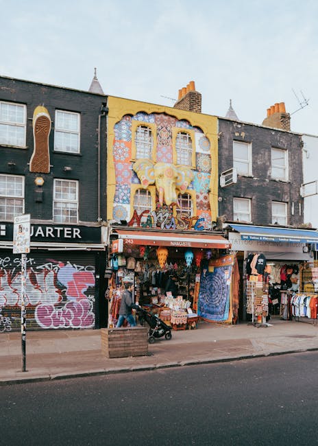 An urban street scene featuring a row of three-story buildings with storefronts on the ground level and residential windows above. The central building has a vibrant, multicolored mural painted on its facade, displaying a yellow bear-like figure with patterns in pink, blue, and white, along with decorative cloud motifs. The ground floor storefronts display various clothing items, accessories, and decorative textiles arranged on racks and shelves, with some hanging outside under awnings. In front of the stores, a person is seen pushing a baby stroller along the sidewalk, which is paved and includes a wooden planter box. Parked to the side is a vehicle, possibly a van used for house removals or furniture transport, nearby. The street has a signpost, and the scene is lit with natural daylight, capturing a typical day of home relocation or furniture transport preparations in an area that may require Camden Council permits, consistent with services provided by Man and Van Kentish Town.