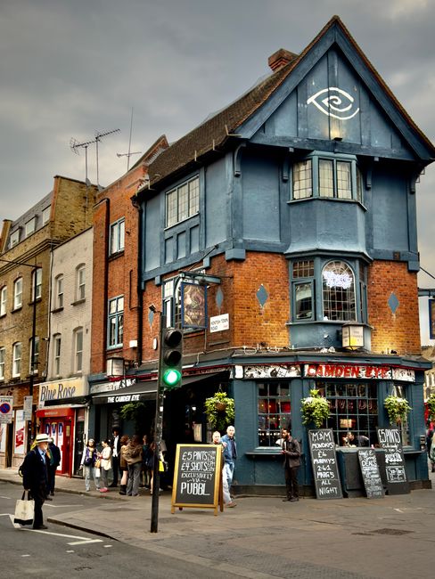A three-story corner building with a blue-painted upper structure and a brick lower section, situated on a busy street in Kentish Town. The ground floor features a pub with large windows adorned with flower boxes and multiple chalkboard signs displaying drink specials and opening hours. The building's façade includes a sign reading 'Camden Café,' and a painted eye symbol is visible on the blue upper section beneath the roof. Several pedestrians are walking and standing around, some near the pub entrance, while others converse near a traffic light showing a green signal. There are parked cars along the street, and a few items such as cardboard boxes and moving blankets are positioned outside, indicating preparations for a home relocation or furniture transport. This scene captures elements of urban moving logistics, with the nearby pavement and road providing space for loading or unloading household items, and the surrounding environment emphasizing a typical London retail and residential area.