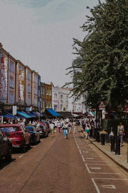 A busy street scene in Kentish Town showing a row of multicoloured terraced houses on the left side, some with commercial storefronts on the ground floor, and an outdoor market with numerous vendors and shoppers in the background. Parked cars line the curb on both sides of the street, with a designated loading and unloading zone marked by numbered parking bays visible in the foreground. On the right, a large leafy tree extends over the pavement, providing shade. The scene captures a typical community area during daytime, with natural lighting highlighting the bustling activity. As part of home relocation or furniture transport operations, a Man and Van Kentish Town professional team might be involved in packing and loading household items into a van, which is potentially visible in the background, ready for a house removal or delivery service in Kentish Town.