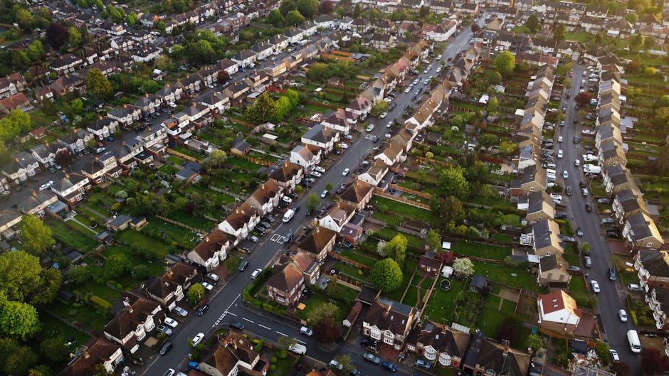 An aerial view of a residential neighbourhood showing multiple rows of terraced and semi-detached houses with tiled roofs, interspersed with green gardens, trees, and driveways. The streets are lined with parked cars and are relatively narrow, typical of suburban areas. Visible from above during daylight with soft, natural lighting, this scene depicts a well-established community suitable for home relocations. The image captures the layout of the streets, the arrangement of houses, and the surrounding greenery, providing context for house removals and furniture transport services offered by Man and Van Kentish Town, especially relevant to their removals cost guide for the Kentish Town area.