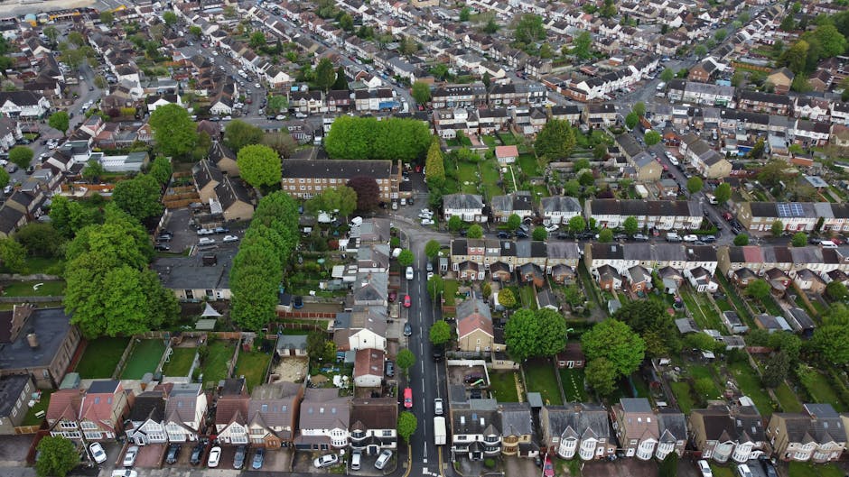 A busy street scene in Kentish Town showing a row of multicoloured terraced houses on the left side, some with commercial storefronts on the ground floor, and an outdoor market with numerous vendors and shoppers in the background. Parked cars line the curb on both sides of the street, with a designated loading and unloading zone marked by numbered parking bays visible in the foreground. On the right, a large leafy tree extends over the pavement, providing shade. The scene captures a typical community area during daytime, with natural lighting highlighting the bustling activity. As part of home relocation or furniture transport operations, a Man and Van Kentish Town professional team might be involved in packing and loading household items into a van, which is potentially visible in the background, ready for a house removal or delivery service in Kentish Town.