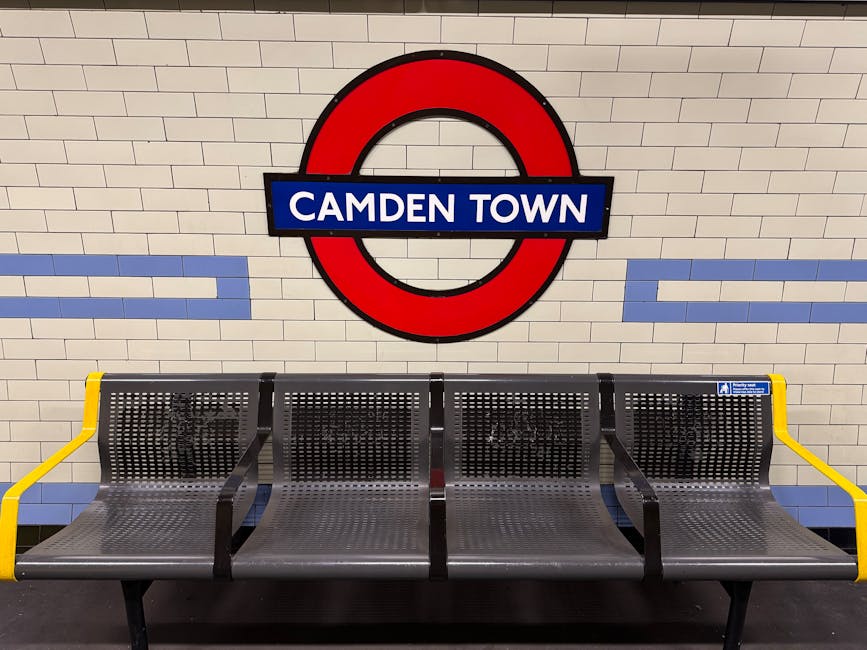 A row of four metal benches with perforated seats and backrests, positioned side by side on a platform at Camden Town underground station. The benches have yellow armrests on both ends, and a small blue and white notice is affixed to the rightmost armrest. Behind the benches, the station’s tiled wall features a large roundel sign with a red outer ring, a blue bar in the middle displaying the text 'CAMDEN TOWN' in white, and light beige rectangular tiles arranged horizontally with blue accents. The lighting is bright and even, illuminating the scene clearly, with no visible luggage or items indicating moving activities, but the setting reflects typical station waiting area layout, relevant for visualising home relocation or furniture transport in the context of removals and moving services by Man and Van Kentish Town.