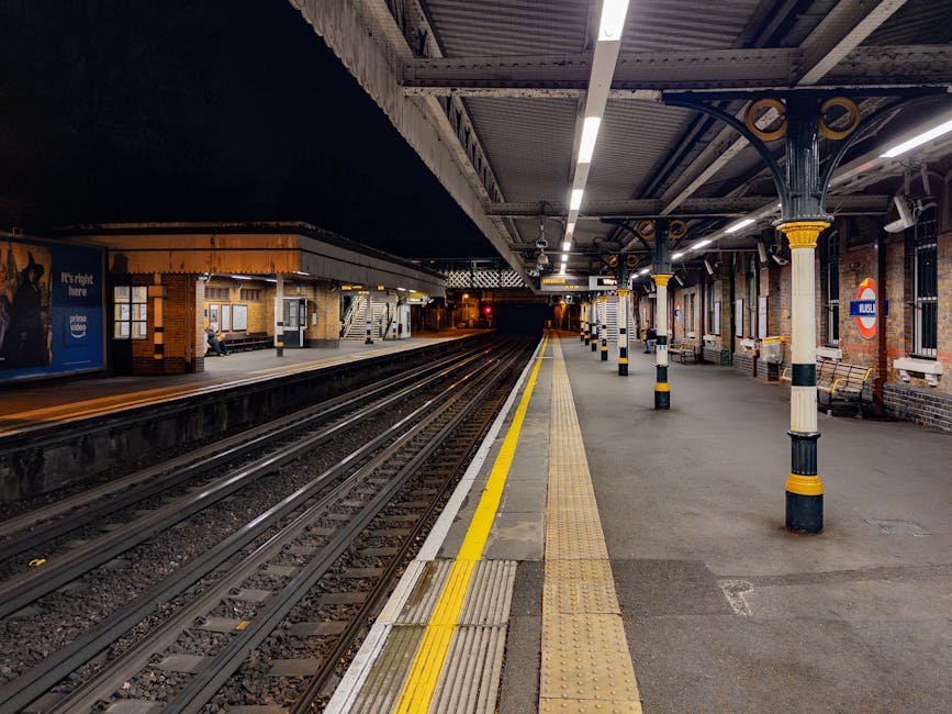A photograph of Kentish Town Station platform at night, showing empty railway tracks, a covered waiting area with brick walls, benches, and informational signage. The platform is illuminated by overhead fluorescent lights, with several yellow safety lines and tactile paving along the edge. The station features black and yellow bollards supporting the canopy structure, which is composed of metal beams and panels. The scene captures the quiet atmosphere of the station during evening hours, with no visible passengers or staff, and the station's entrance in the background. This setting illustrates a typical environment for house removals or furniture transport, where careful loading and unloading occur in a controlled, sheltered space, often managed by services such as Man and Van Kentish Town.