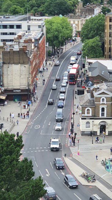An aerial view of a busy urban street scene with multiple lanes of moving vehicles, including cars and buses, parked and traveling along the road near a corner. The street is lined with a mix of modern and historic buildings, some with brick facades, and several trees providing greenery along the sidewalks. Pedestrians are visible walking on the pavements, and bicycles are parked near a small, white, classical-style building situated at the intersection. In the background, more trees and the tops of taller buildings are visible, indicating a well-developed city environment. This scene, typical of an area served by house removal companies like Man and Van Kentish Town, illustrates the urban logistics involved in furniture transport and home relocation activities within the city.