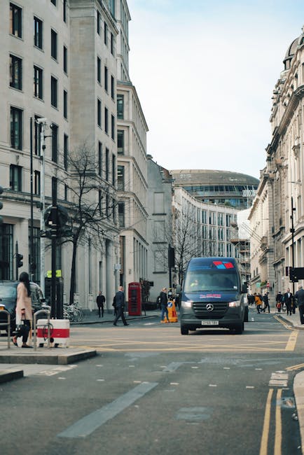 A city street scene during daylight showing a medium-sized grey van, parked partly on the pavement and street, with its front facing the camera. The van's rear doors are open, revealing a glimpse of packed cardboard boxes, plastic wrapping, and furniture items inside, indicating it is in the process of loading or unloading for a house relocation. Surrounding the van are pedestrians walking on the pavement, some standing near the curb, and others crossing the street. The background features tall, older-style stone buildings with multiple windows and decorative architectural details, as well as a modern multi-storey building further down the street. Street furniture includes a red telephone box, street lamps, and yellow safety cones. The scene is well-lit with natural daylight, capturing the typical urban environment associated with moving services provided by companies like Man and Van Kentish Town, highlighting the logistics involved in furniture transport and home removals. The overall setting emphasizes a busy, professional moving process in an urban residential area.