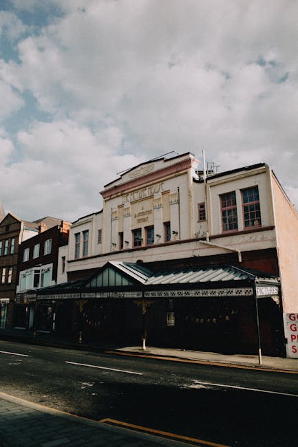 Photograph of a multi-story building with a classic architectural style situated on a street, taken during the daytime with partly cloudy skies. The building features a cream-colored facade with decorative elements, large windows on the upper floors, and a prominent sign indicating it as a historic or entertainment venue. The ground level has a dark storefront, partially obscured by a black awning with gold trim, and a small sign displaying the word 'PICTURES.' The street in front includes a pavement and an asphalt road with visible double yellow lines, suggesting a busy urban area. There are no visible vehicles or pedestrians in the scene. This image captures the exterior environment of a central location that might be involved in house removals or relocation logistics, as part of moving services such as those offered by Man and Van Kentish Town. The building's setup and street context can be relevant for planning a home relocation or furniture transport to or from nearby properties.