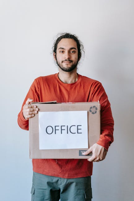 A man with dark, curly hair and a beard, wearing a red long-sleeve shirt and grey trousers, stands indoors against a plain light grey wall. He holds a large cardboard box with a white label that reads