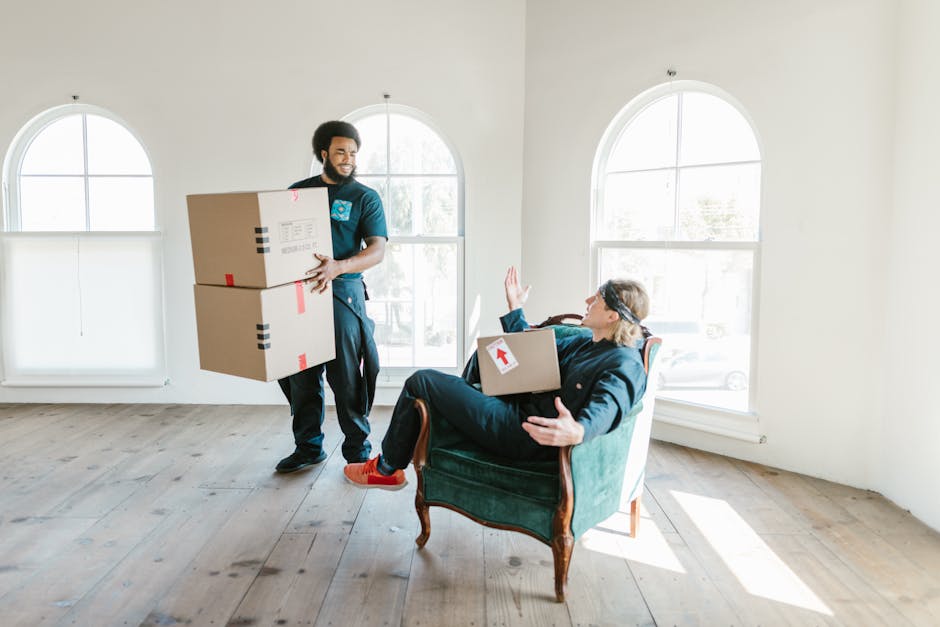 A man and woman inside a bright, modern living room preparing for a house move. The man, wearing an orange pair of trousers and a maroon t-shirt, is smiling while holding a small cardboard box labeled