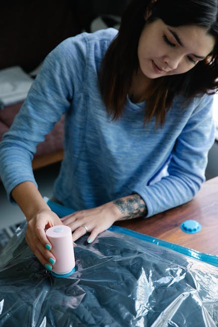 A woman with long dark hair, wearing a light blue long-sleeved top, is seen sealing a clear plastic bag with a pink handheld sealing device on a wooden table. The table has packing materials, including a blue plastic cap and other items. The woman is focused on the packing process, which is part of preparing items for a house relocation or moving service. Inside the background, there is a sofa with visible cushions, indicating a home environment. The scene emphasizes packing and moving logistics, consistent with the services offered by Man and Van Kentish Town for home removals and furniture transport, especially during challenging access situations like those described on the 'Pond Street removals access problems and solutions' page.