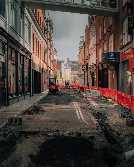 A city street under construction with an uneven, partially excavated road surface, surrounded by multi-storey brick and stone buildings on both sides. The road is lined with orange safety barriers and concrete blocks, with construction equipment such as small excavators visible further in the distance. A pedestrian bridge spans overhead, and signage indicates parking facilities nearby. The scene is set in an urban area likely undergoing infrastructure upgrades, which could impact access for house removals or furniture transport. The image's context relates to logistical challenges faced during home relocations in the Kentish Town area, as handled by Man and Van Kentish Town, highlighting the importance of planning for access problems during moving services.