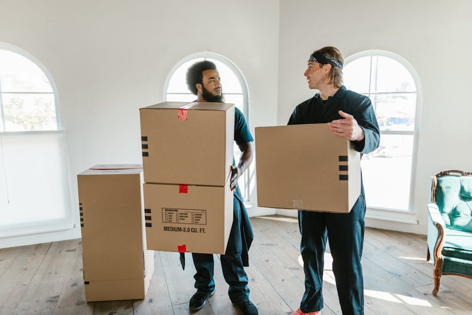 Two men involved in a home relocation are inside a well-lit room with wooden flooring and large arched windows, which allow natural light to fill the space. One man with dark hair and a beard, dressed in casual dark clothing, is carrying a cardboard box labeled 'medium-3.0 cu. ft.' with red tape securing the top flaps, while the other man, with lighter hair and wearing a headband and dark attire, is holding a similar-sized cardboard box in front of him. Several additional cardboard boxes, some stacked and others standing alone, are visible on the floor nearby, indicating an ongoing packing and moving process. The scene appears to capture a moment during a furniture transport or loading process, likely part of a house removal service provided by Man and Van Kentish Town, as they prepare to load items into a van or transport vehicle outside or inside the property.